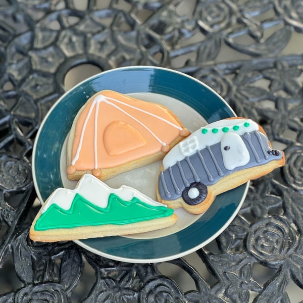 Three camping-themed cookies, including a tent, camper, and mountains, on a blue and white plate.