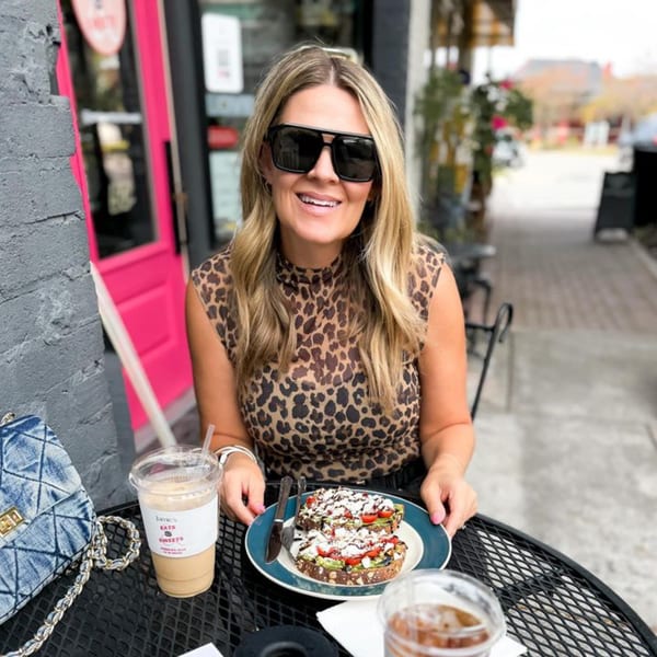 A smiling woman in sunglasses at Jamie’s Eats and Sweets, holding a plate of avocado toast.