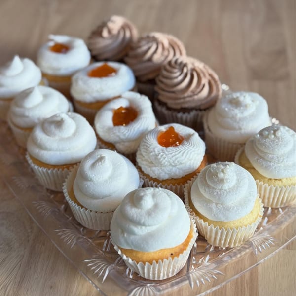 A clear tray holds rows of cupcakes with white and chocolate frosting, some with orange jam centers.
