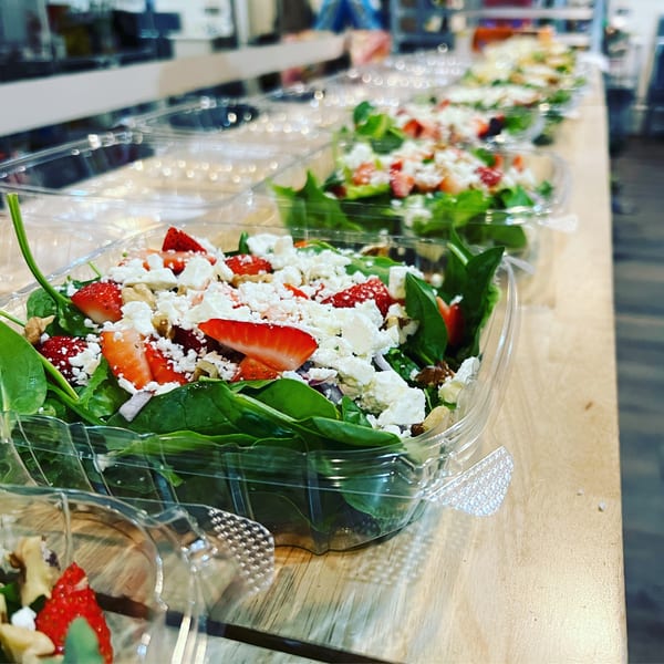 Rows of fresh spinach salads with strawberries, feta, and walnuts in clear to-go containers.