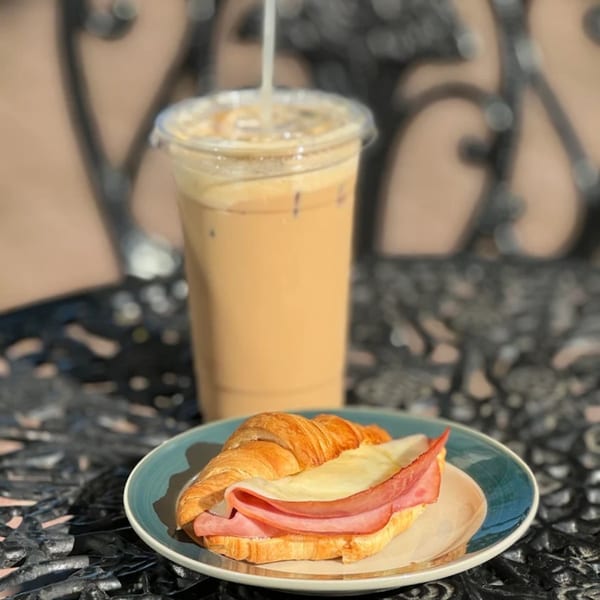 A ham and cheese croissant on a blue plate next to an iced coffee on a dark patterned table.