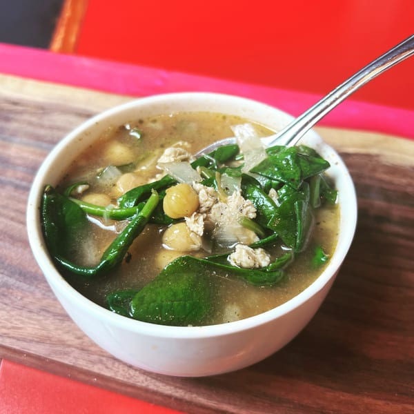 A white bowl of Jamie’s Eats and Sweets soup with spinach, chickpeas, and meat, with a spoon.