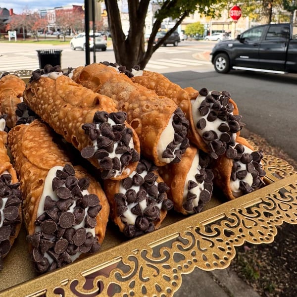 A golden tray filled with cannolis topped with white cream and chocolate chips, with a street in the background.